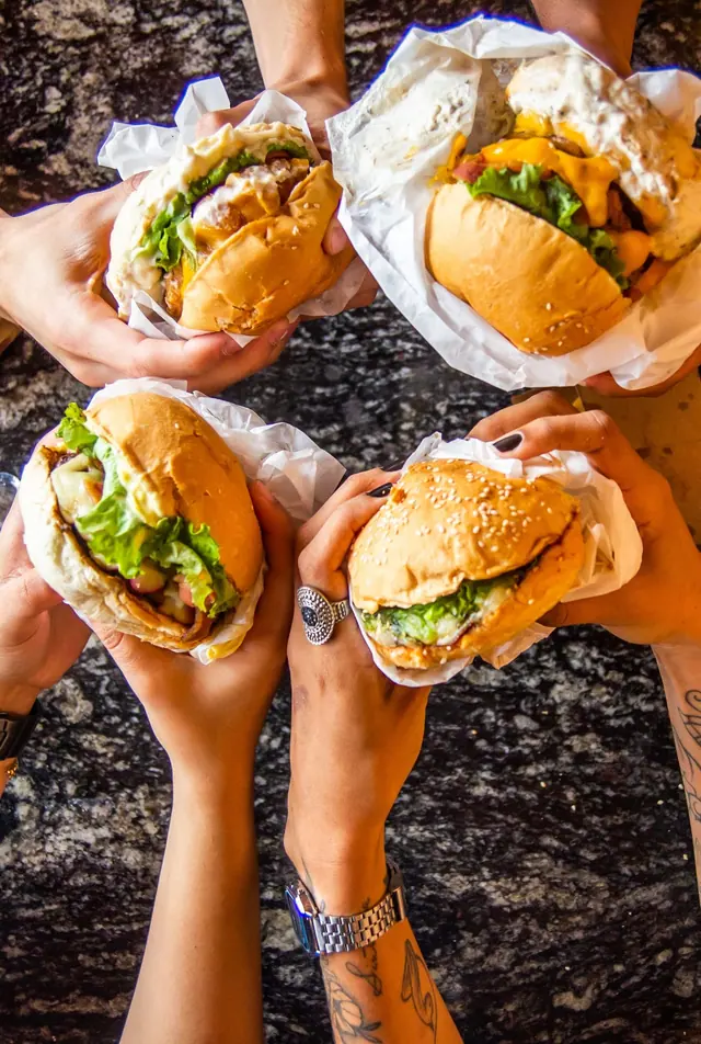 Four hands holding up burgers wrapped in paper over a dark table